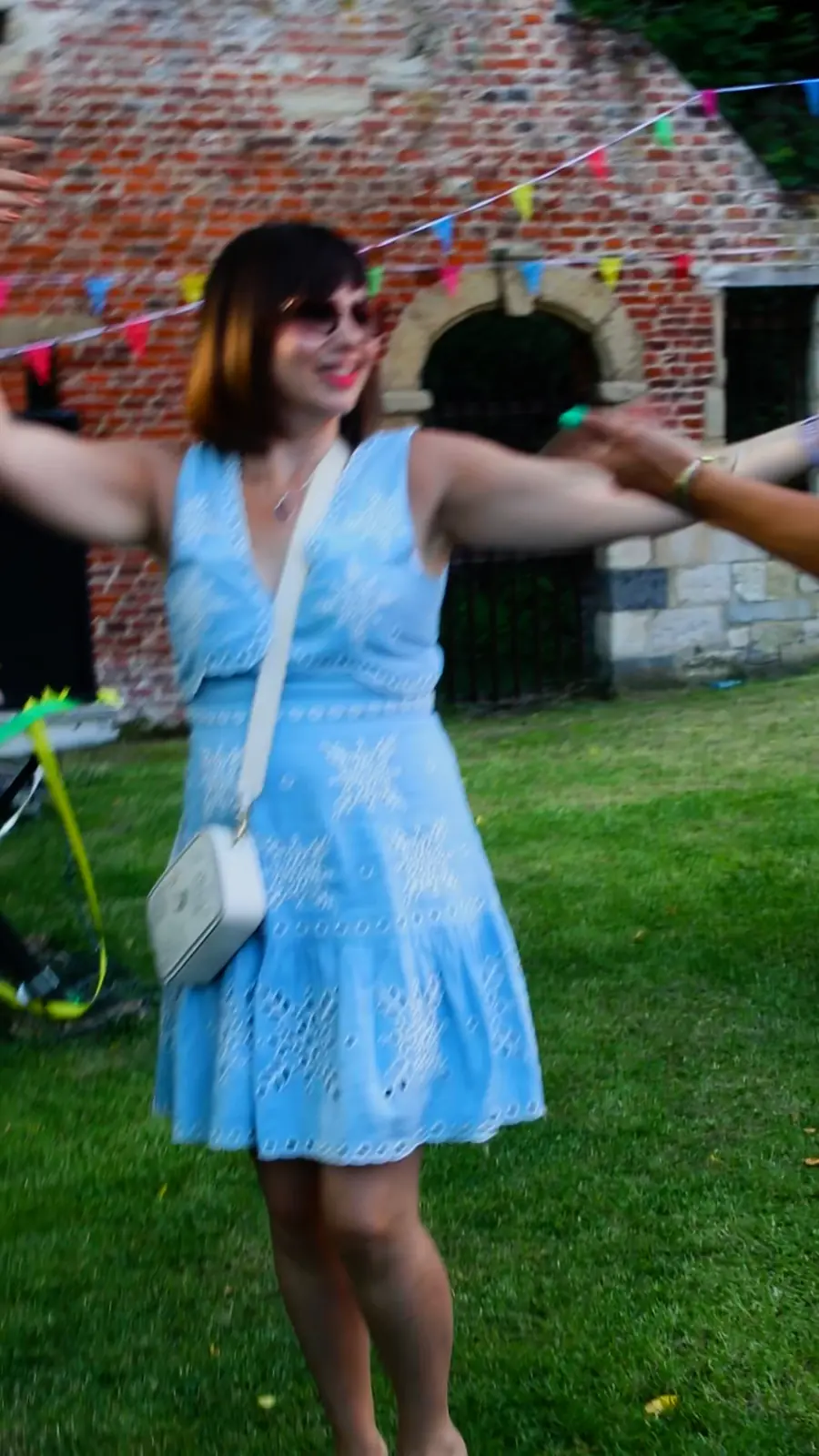 Guests dancing on a lawn between old brick walls and bunting at Nava's Tirgan festival, with musicians, art and grills, and a view of the park from above.