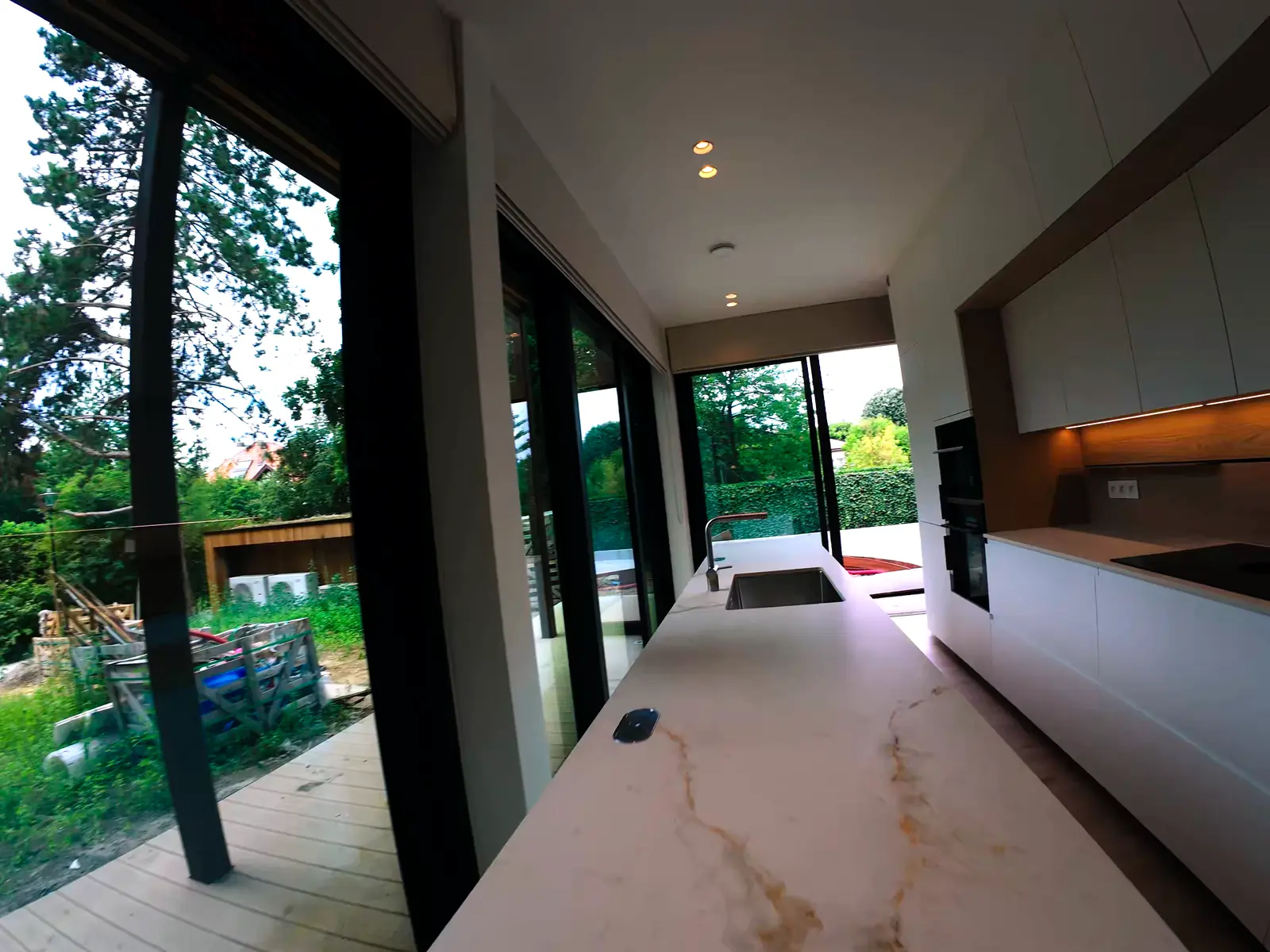 A flythrough of an empty modern house: the open kitchen with a marble island and glass doors onto the garden, then a rise above the hedge and the rooftops to the Oralis Real Estate logo.